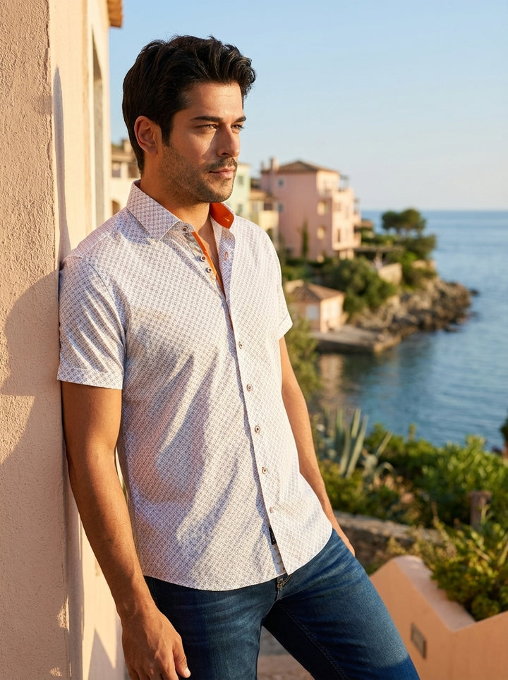 A refined men's short-sleeve button-down shirt featuring a white base with a sophisticated geometric micro-print. The design includes contrasting orange interior collar detailing and decorative buttons, paired with dark wash denim for a modern, polished s