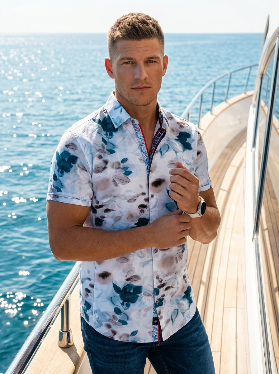 A modern men's short-sleeve button-down shirt featuring a sophisticated watercolor floral print in shades of teal, indigo, and muted grey on a white base. It includes contrasting red and blue patterned inner placket details for a refined, contemporary loo