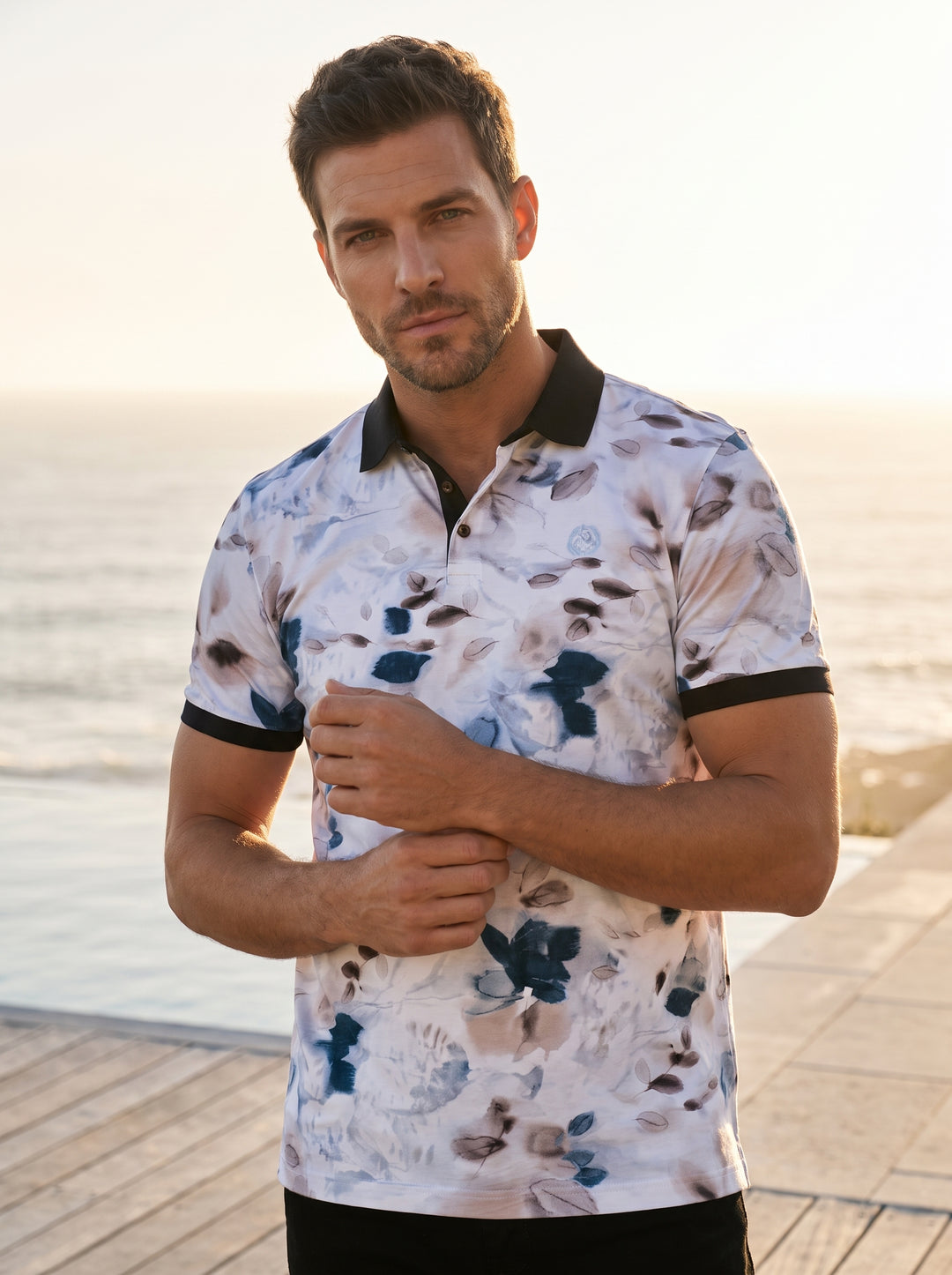 This stylish short-sleeved polo shirt features an artistic watercolor floral print in shades of blue, grey, and soft brown on a white base. It is accented with a contrasting black collar and sleeve cuffs for a modern, tailored look.