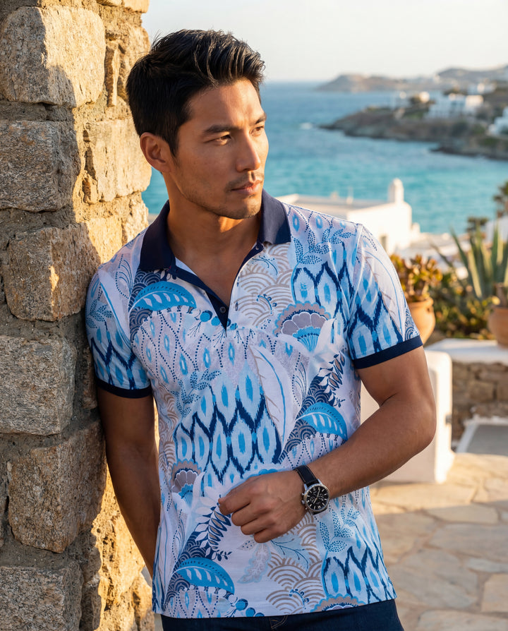 This stylish short-sleeve polo shirt features a vibrant blue and white ikat and floral pattern with a dark navy collar and cuff trim. Crafted for a modern fit, it blends eclectic Mediterranean-inspired motifs for a sophisticated yet casual summer look.