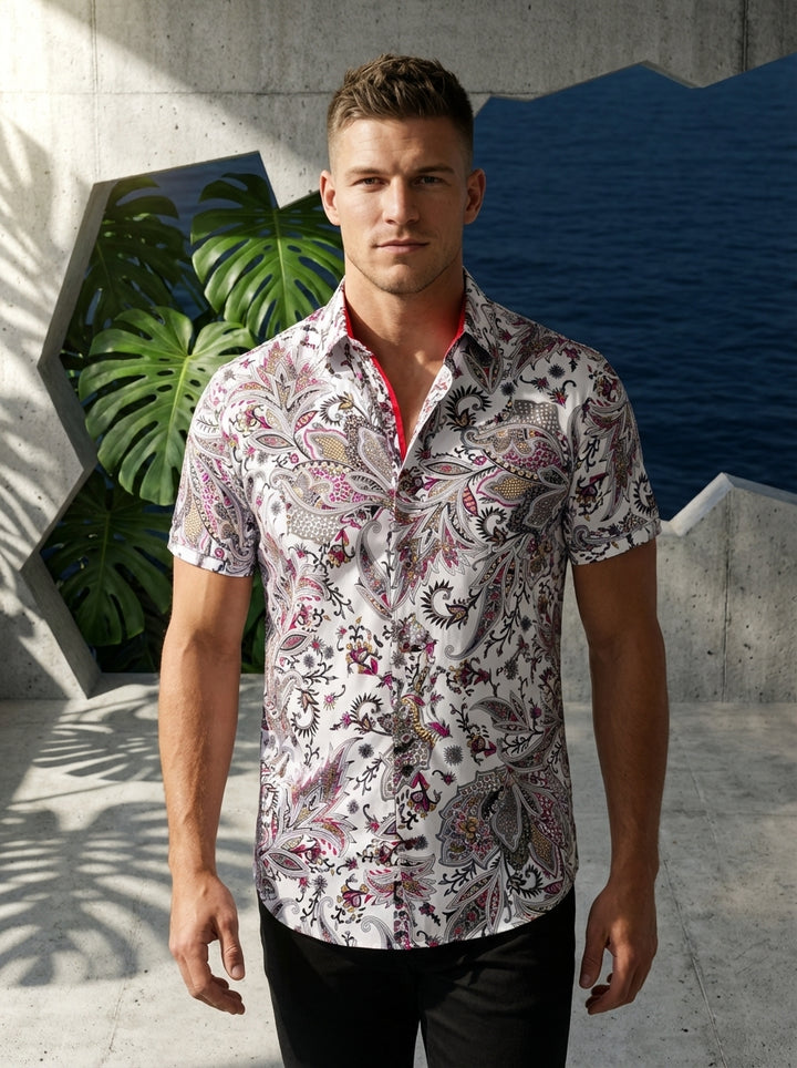 This men's short-sleeved button-down shirt features an intricate, oversized paisley and floral pattern in vibrant shades of magenta, gold, and grey over a crisp white base. It includes a contrast red inner collar stand for a bold, modern touch.