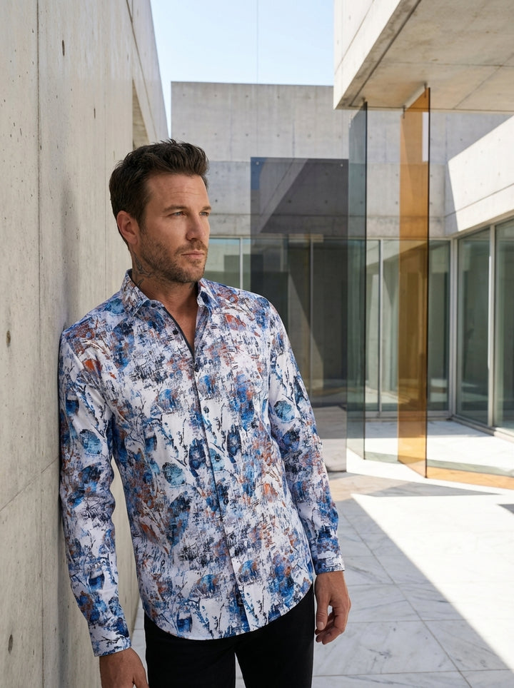 This men's long-sleeve button-down shirt features a vibrant, abstract paint-splatter print in shades of azure blue, burnt orange, and charcoal gray over a crisp white base. Crafted with a modern tailored fit, it includes a classic pointed collar.