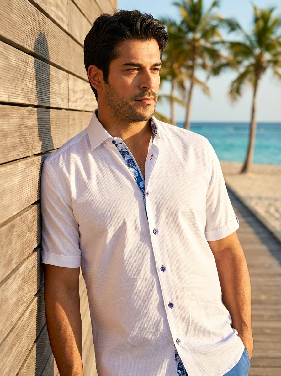 This light white short-sleeved button-down shirt features a subtle textured weave and vibrant blue floral contrast trim on the inner collar and placket. The design is finished with distinctive blue and red buttons, offering a sophisticated yet relaxed sum