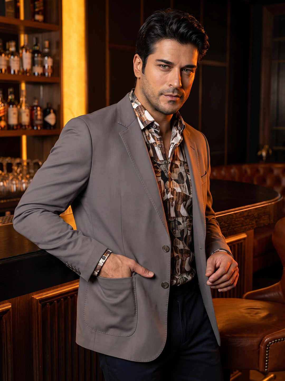 This tailored grey blazer features a modern two-button closure, notched lapels, and spacious patch pockets for a relaxed yet refined look. It is layered over a bold, earth-toned geometric print shirt, creating a sophisticated ensemble for the modern gentl