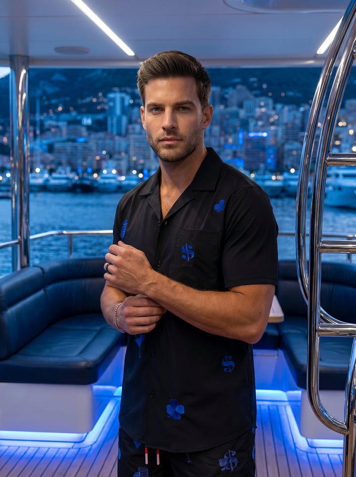This sleek black short-sleeve button-down shirt features vibrant blue embroidered motifs including dollar signs, cocktails, and gambling icons. Made from a smooth, lightweight fabric, it includes a classic camp collar and a single chest pocket for a moder