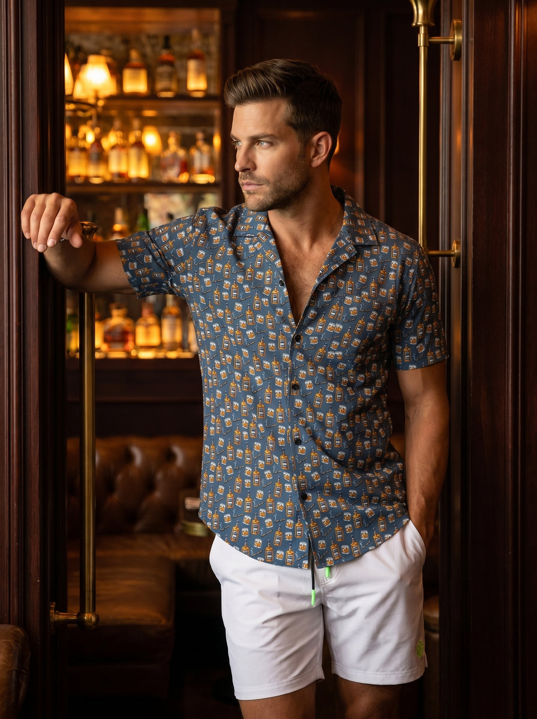 This tailored short-sleeve button-down shirt features a slate blue base adorned with a playful repeat pattern of whiskey bottles, tumblers, and cigars. Crafted for comfort, it includes a modern camp collar and black buttons, perfect for making a statement