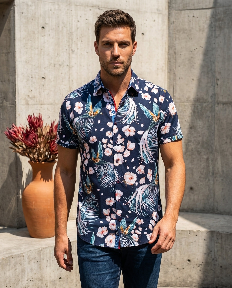 This navy short-sleeved button-down shirt features a vibrant tropical print with hummingbirds, palm fronds, and hibiscus flowers. The piece is accented with contrast red stitching and a patterned inner collar, offering a bold, sophisticated take on a clas