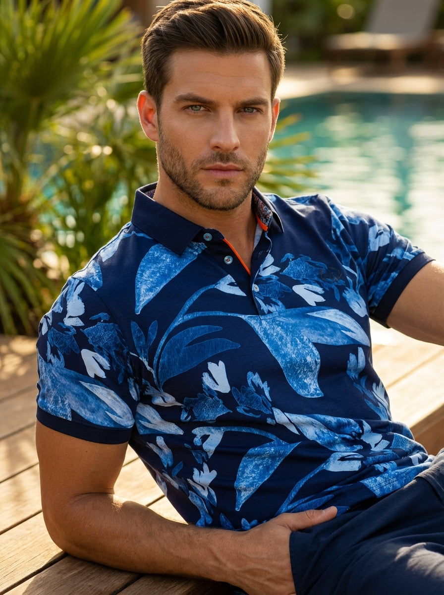 This navy polo shirt features a vibrant blue floral leaf print with textured artistic details. It includes a solid navy collar, a three-button placket with contrasting light blue buttons, and a pops of bright orange along the inner neck lining.