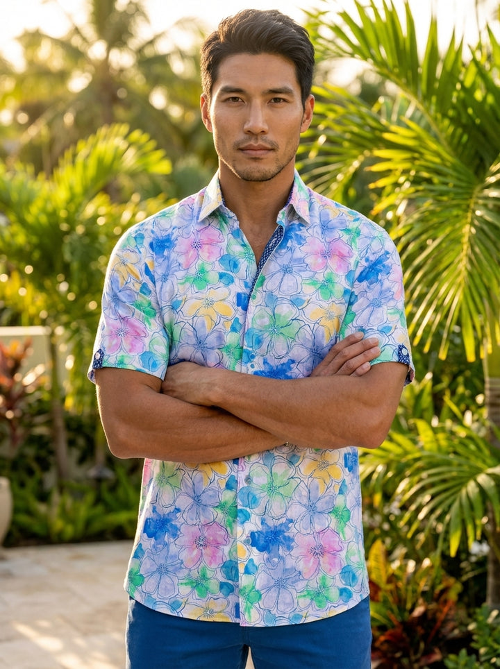 This vibrant men's short-sleeve button-down shirt features a playful watercolor floral pattern in shades of blue, pink, green, and yellow. Crafted with a tailored fit, it includes a contrast geometric print on the inner collar and sleeve cuff tabs.