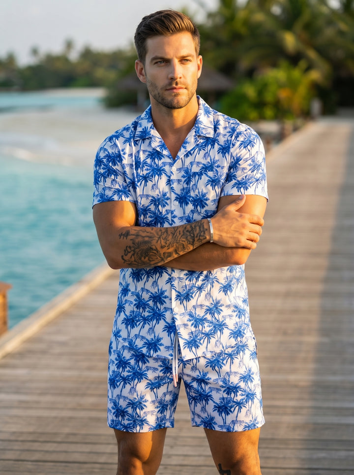 This matching two-piece set features a vibrant blue and white tropical print with palm trees and tiger motifs. The lightweight, button-down short-sleeve shirt and coordinating mid-thigh shorts offer a relaxed, cohesive summer look perfect for warm weather