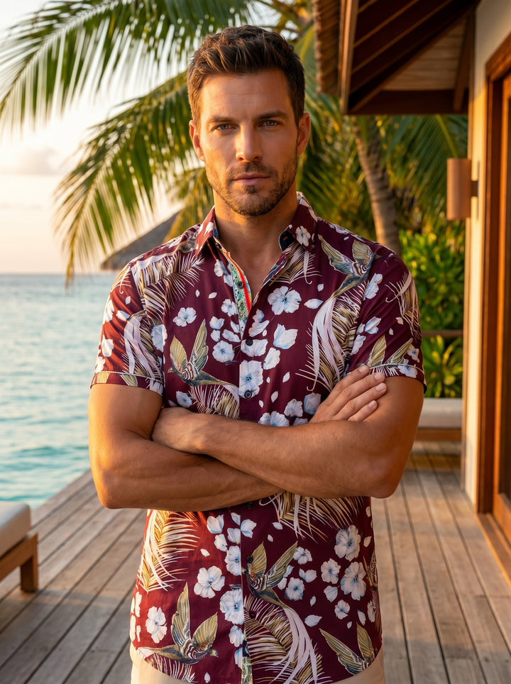 This men's short-sleeve button-down features a rich burgundy base adorned with a tropical motif of white hibiscus flowers, golden palm fronds, and stylized birds. It includes a refined multi-colored inner collar detail for a modern, sophisticated aestheti