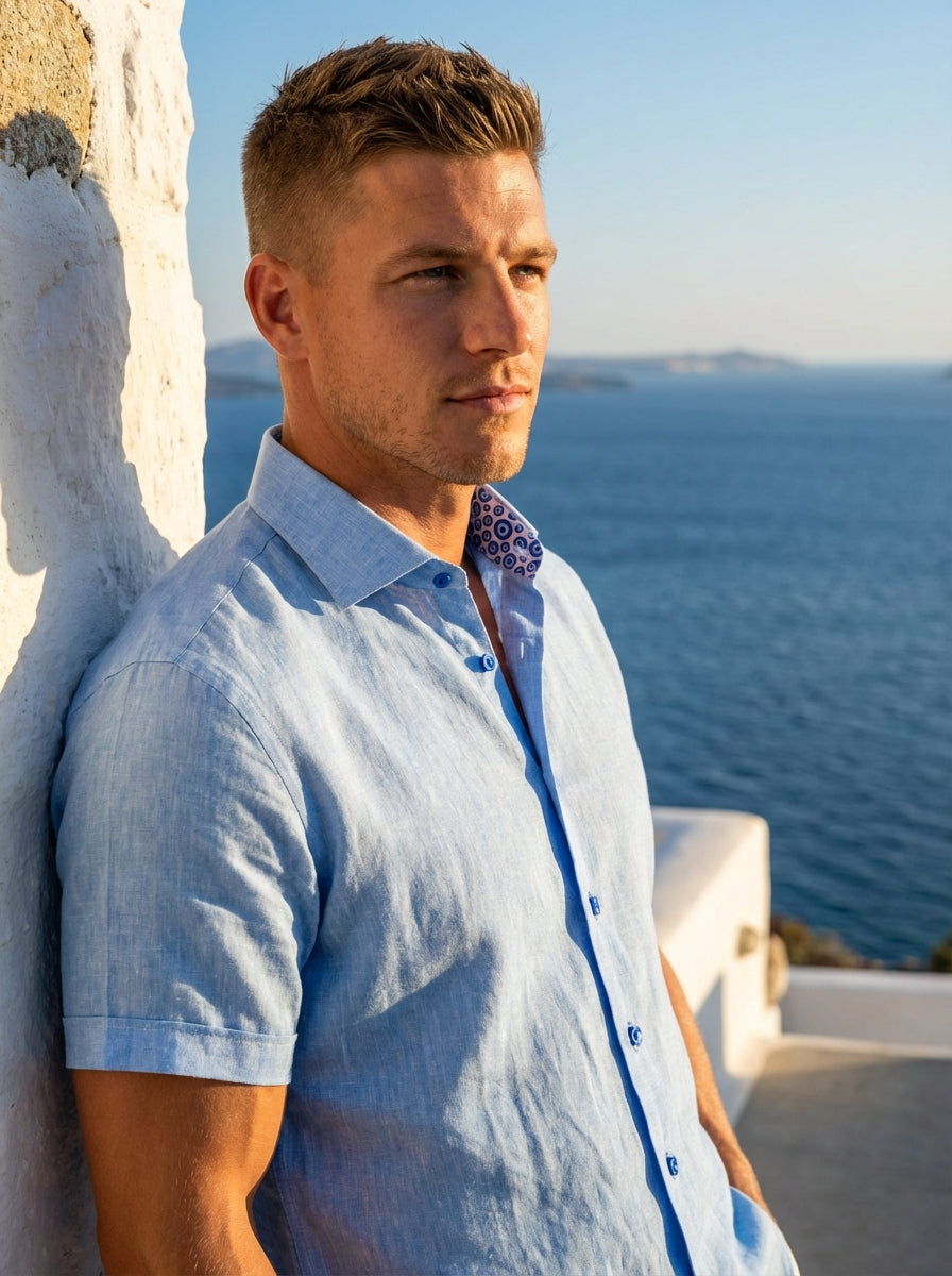 This light blue, short-sleeve button-down shirt by Masutto features a breathable linen-blend texture. It is distinguished by vibrant cobalt blue buttons and a hidden evil eye print along the inner collar stand, offering a sophisticated yet playful Mediter