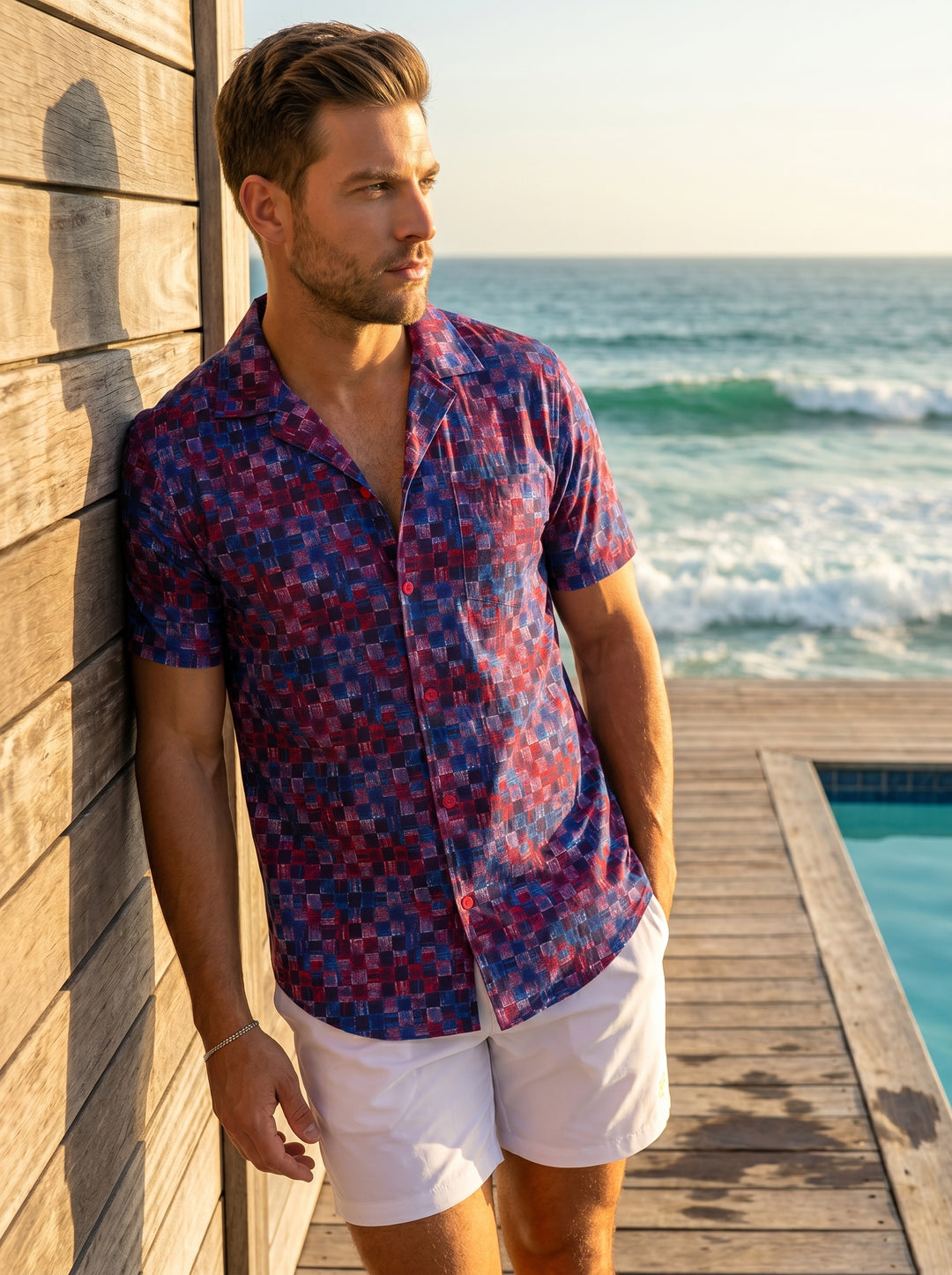 This men's short-sleeve button-up features a vibrant, textured checkerboard pattern in deep blue, red, and purple hues. With a casual camp collar and contrasting red buttons, it offers a modern, relaxed fit perfect for standing out in warm weather.