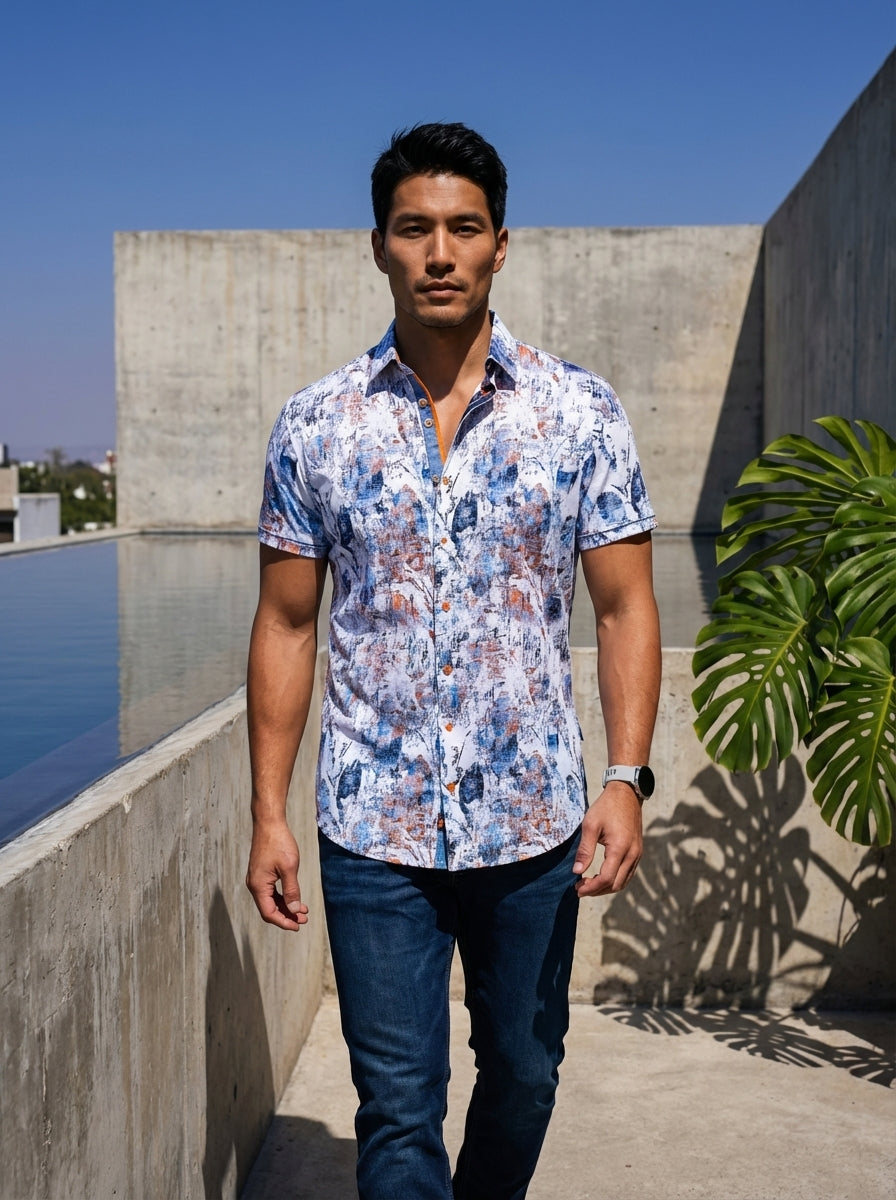 This men's short-sleeve button-down shirt features a vibrant abstract print in shades of blue and copper on a crisp white base. It includes a contrasting inner placket with orange detailing, classic collar, and is paired with dark wash denim.