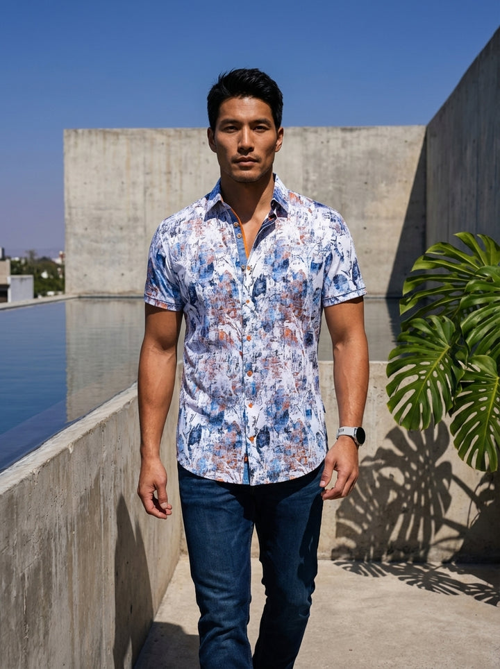 This men's short-sleeve button-down shirt features a vibrant abstract print in shades of blue and copper on a crisp white base. It includes a contrasting inner placket with orange detailing, classic collar, and is paired with dark wash denim.