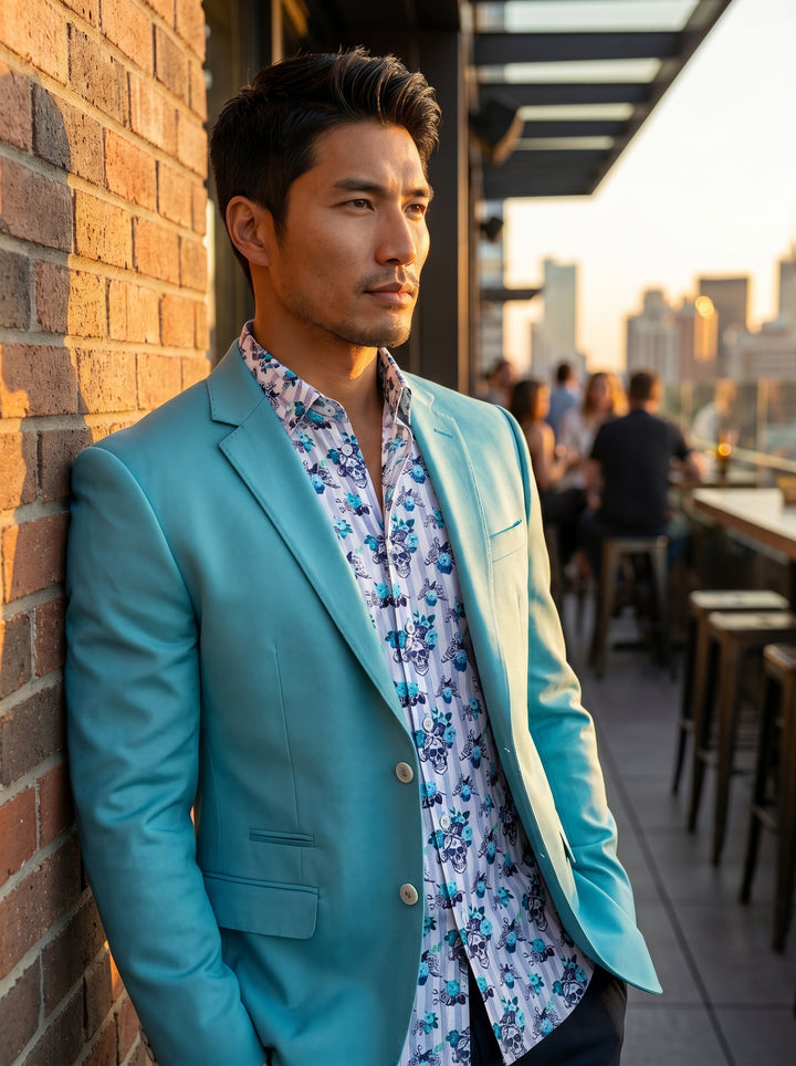 A vibrant turquoise two-button blazer with a notch lapel and flap pockets, layered over a white button-down shirt featuring blue floral and skull motifs. The ensemble offers a bold, contemporary take on smart-casual attire with high-quality tailoring and 
