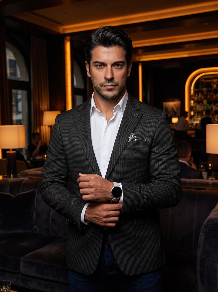 This tailored charcoal textured blazer features a subtle micro-pattern, notched lapels, and a single-button closure. It is paired with a crisp white button-down shirt and a patterned pocket square, offering a sophisticated smart-casual aesthetic for versa