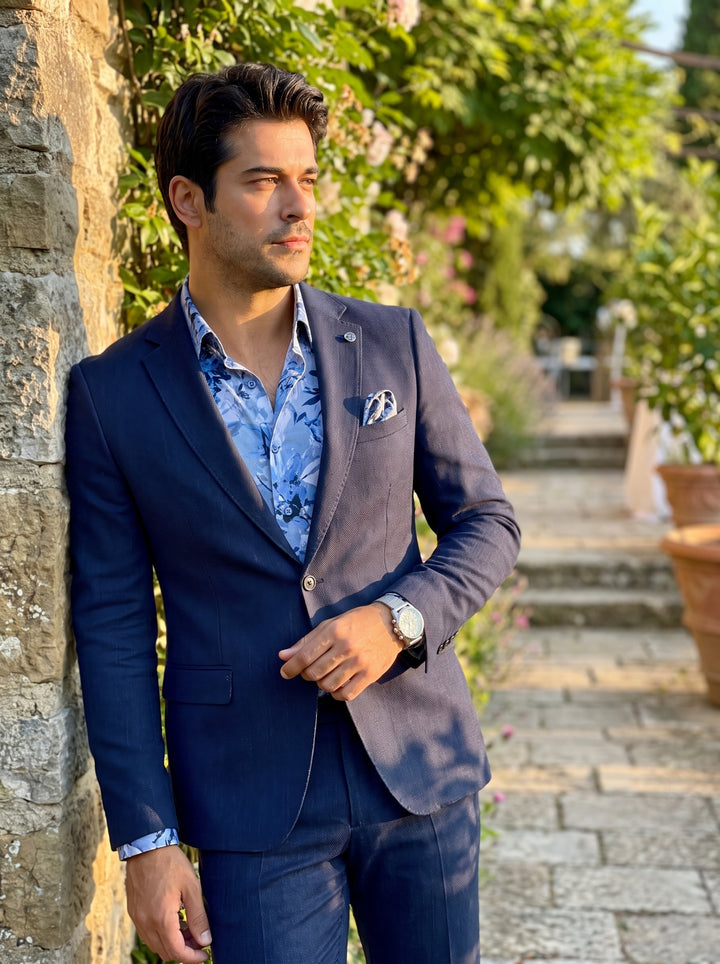 A refined navy blue slim-fit suit crafted from textured fabric. It features notch lapels, a floral print button-down shirt, a matching pocket square, and a silver watch. This ensemble combines classic tailoring with contemporary, vibrant patterns for a mo