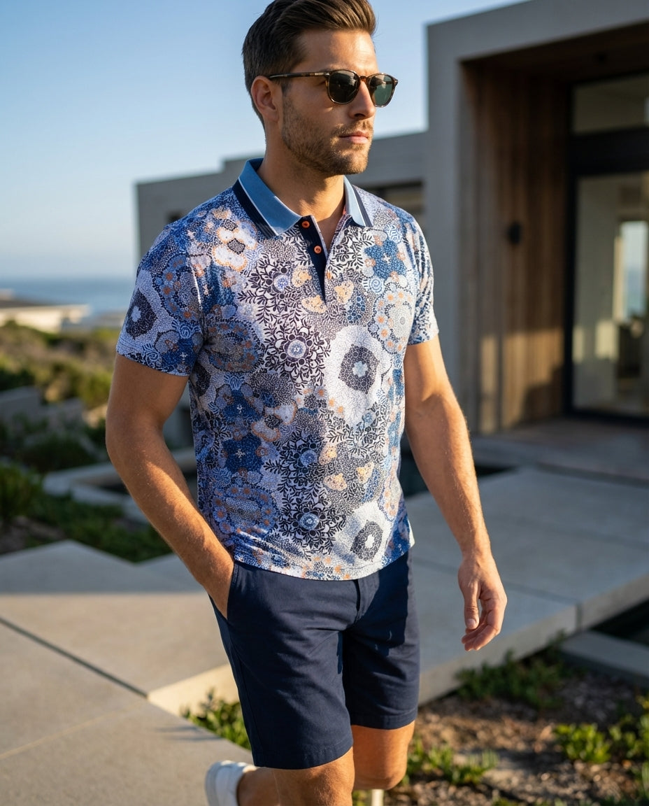 This men's polo shirt features an intricate, multidimensional floral and geometric mandala pattern in shades of blue, navy, and white with vibrant orange accents. It includes a structured navy and sky-blue tipped collar and a three-button placket with ora