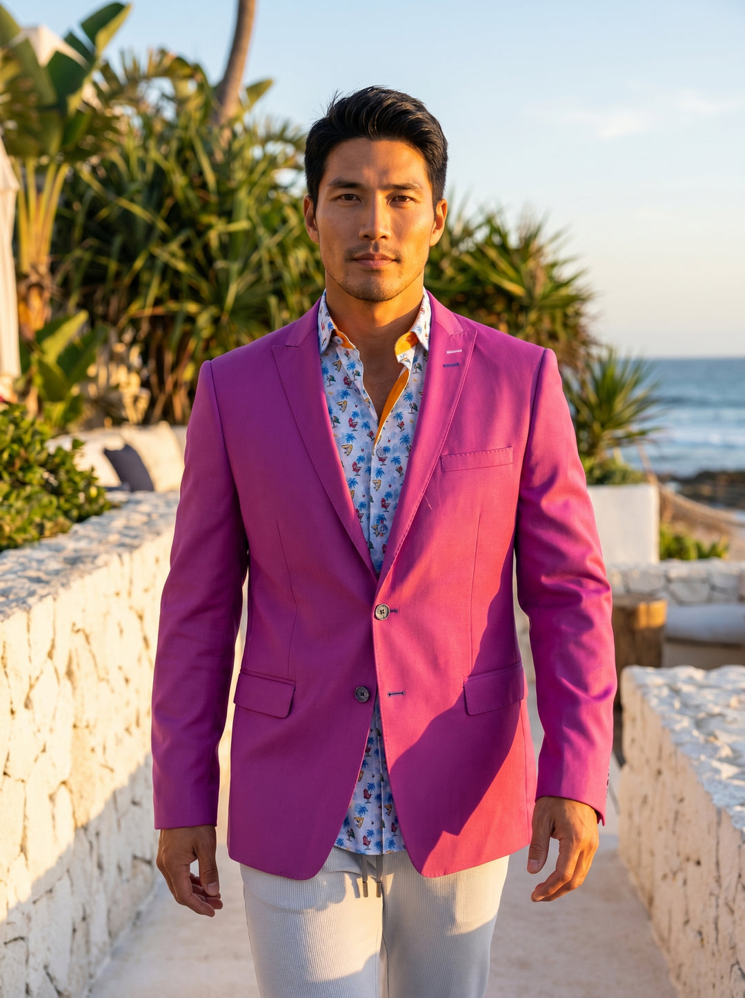 This vibrant magenta blazer features a slim-fit silhouette with notched lapels and decorative button cuffs. It is paired with a tropical-print button-down shirt and light grey trousers, creating a bold, fashion-forward ensemble that balances tailored soph