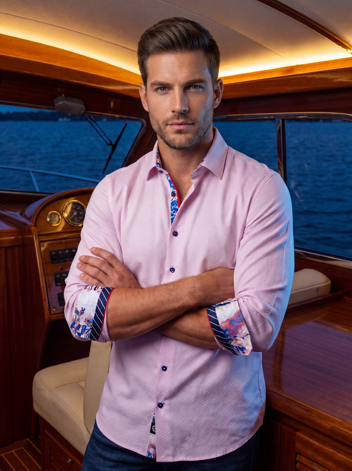 A refined men's button-down shirt in a soft pale pink hue, featuring a subtle micro-textured weave. It is accented with vibrant floral and striped patterns on the interior collar and cuffs, complemented by contrasting dark buttons and red piping.