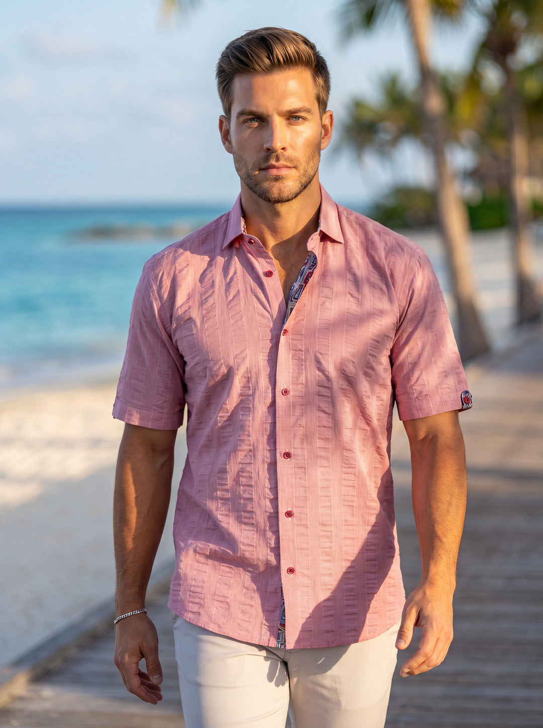 This men's short-sleeved button-down shirt in a vibrant dusty rose features a unique vertical seersucker-inspired pleated texture. It is accented with bold red buttons and patterned contrast trim along the inner collar and cuffs for a sophisticated, relax